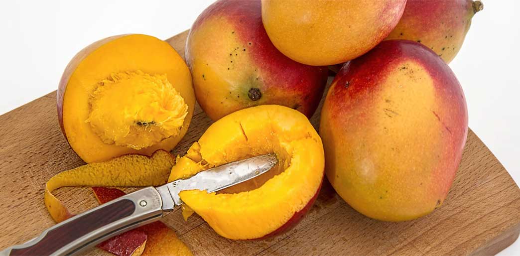 Cutting up Mango with fruit piled onto a wooden chopping board.